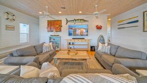 A living room with two grey sofas, a wooden coffee table, and a TV on a wooden stand. There are ceiling fans, a life guard sign, and a fish painting on the wall. - Best Little Shore House by Avantstay Fishing Pond Views + Fire Pit + Hammock Lounge + Pool (Port Aransas)