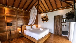 A wooden-framed bed with a canopy, a bedside table with a lamp, a white wall with a wooden art piece, and a sliding barn-style door. - Bali De Pardis Cottage (Munduk)