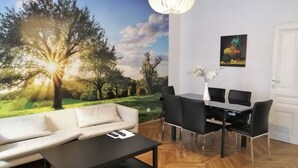 A room with a sofa, a dining table with chairs, a coffee table, a wall mural of a landscape, and a hanging light fixture.
