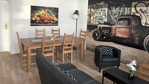 A dining area with a wooden table and chairs, a black sofa, a lamp, and a wall mural of a vintage car and graffiti. - Cozy 2 bedroom Apart with balcony in Ottakring (Vienna)
