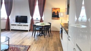 Un salon moderne doté d’un téléviseur à écran plat, d’une table à manger avec des chaises et d’un coin cuisine avec des placards et une cuisinière.