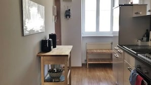 A narrow kitchen with wooden flooring, a black rug, a window with blinds, and a refrigerator. - Home Apartment near Huberpark (Vienna)