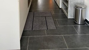 Una cocina estrecha con armarios blancos, un fregadero y una estufa. Hay un cubo de basura y un banco en la esquina. El piso está cubierto de baldosas, y hay una puerta que conduce a una sala de estar con un sofá y una lámpara.