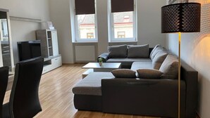 Una sala de estar moderna con un sofá modular, una mesa de centro y una lámpara de pie. Hay dos ventanas con persianas, un radiador y un televisor sobre un mueble.