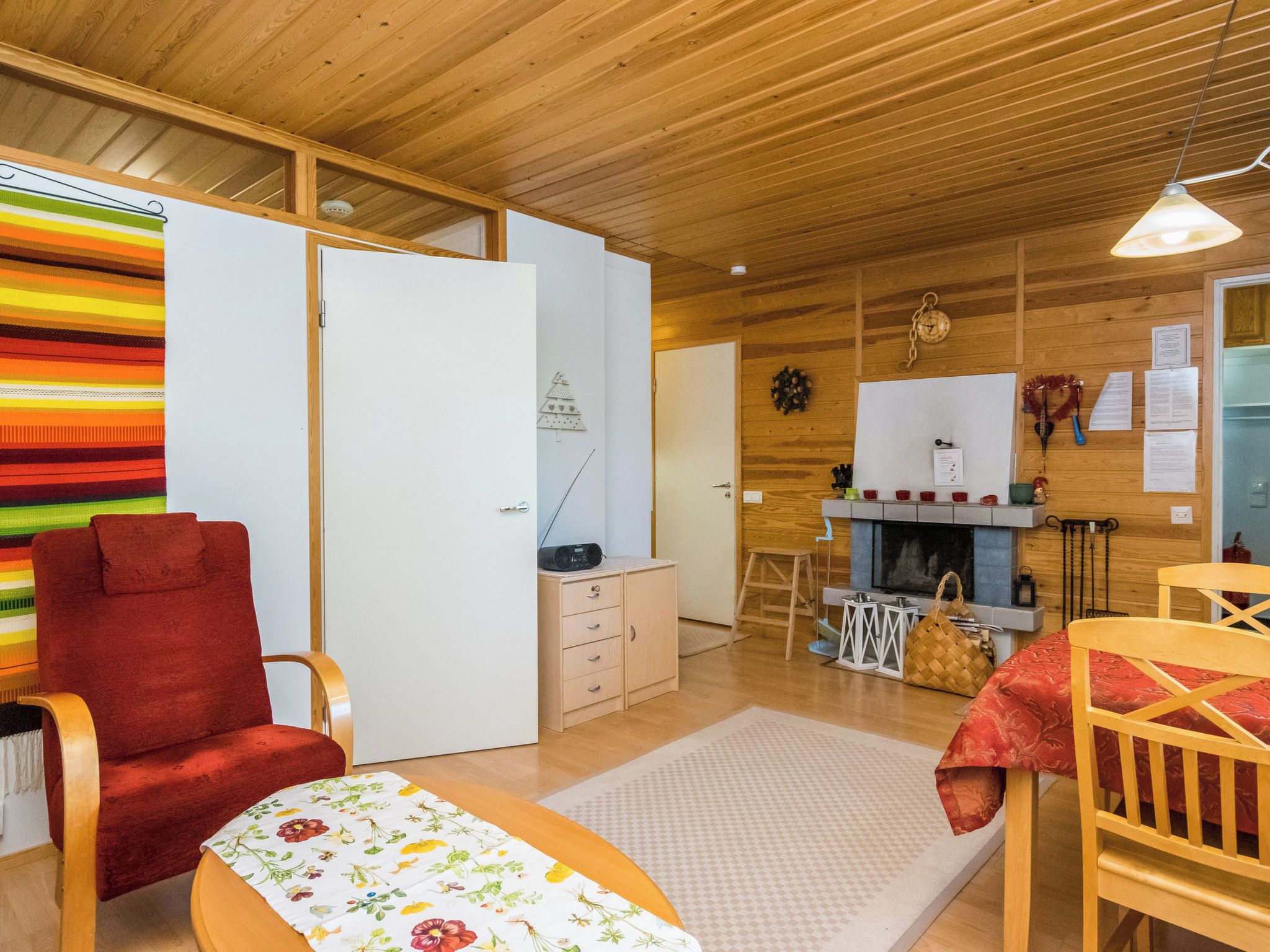 A cozy room with a wooden ceiling, a red chair, a small table with a floral tablecloth, a white wardrobe, and a kitchen area with a stove and a hanging light fixture.