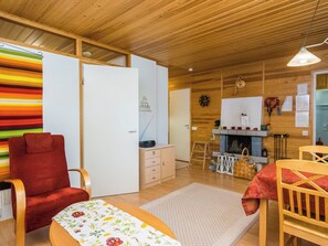 Una habitación acogedora con techo de madera, una silla roja, una mesita con mantel floral, un armario blanco y una zona de cocina con estufa y lámpara colgante.