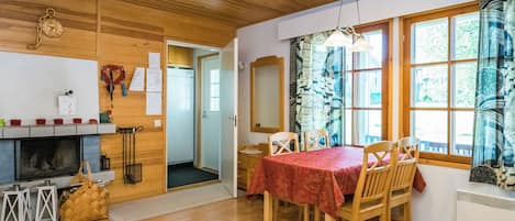Ein gemütliches Zimmer mit Holzbalkendecke, Kamin, Esstisch mit Stühlen und einem Fenster mit gemusterten Vorhängen.