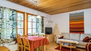 A cozy living room with a wooden ceiling, a dining area with a table and chairs, a TV, a bookshelf, and a sofa with patterned cushions.
