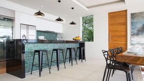 A modern kitchen with a central island, bar stools, and a dining area with a wooden table and chairs. - Zest 4 Bed : Ultra-Luxurious Five Star Villa (Koh Samui)