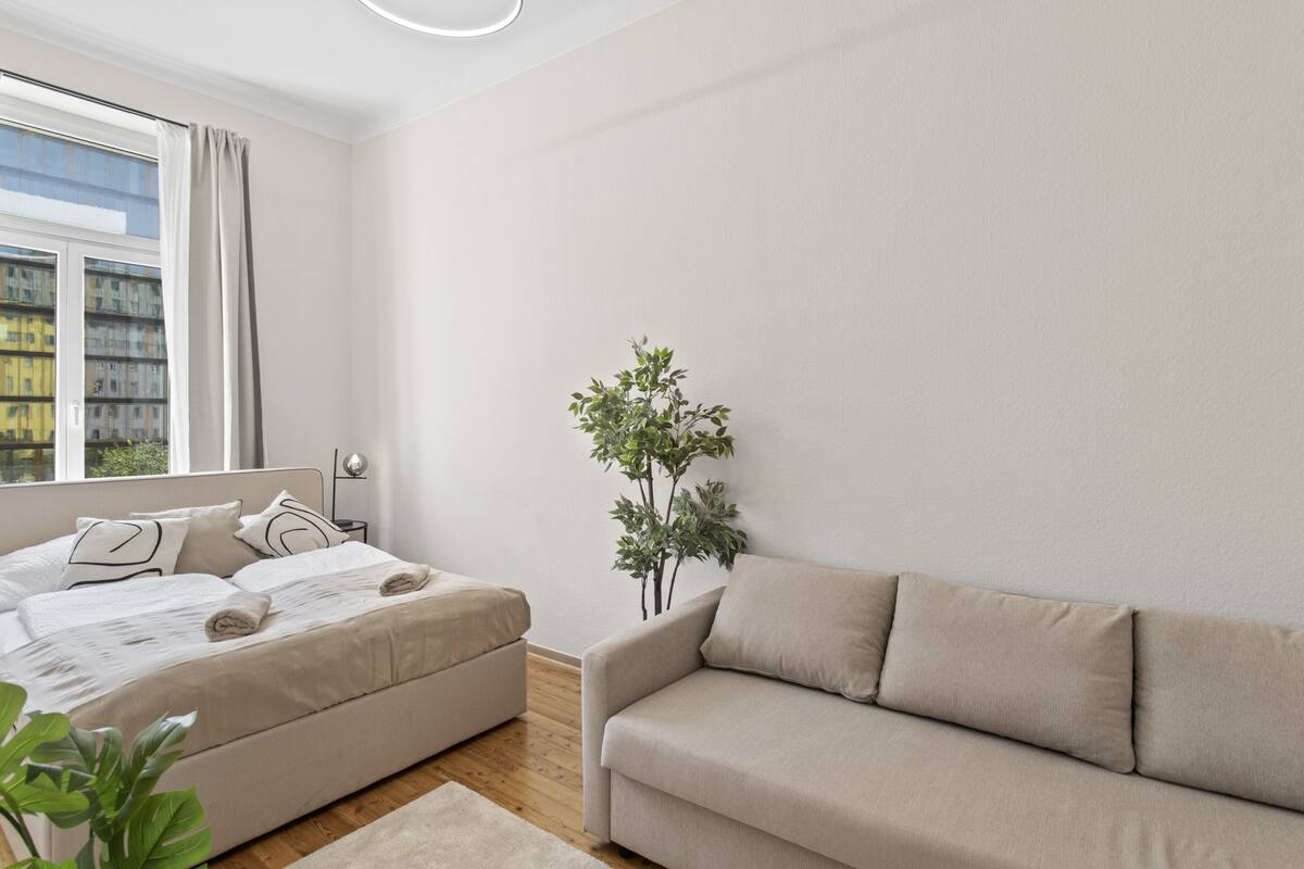A modern bedroom with a bed, a sofa, a potted plant, and a window with curtains.