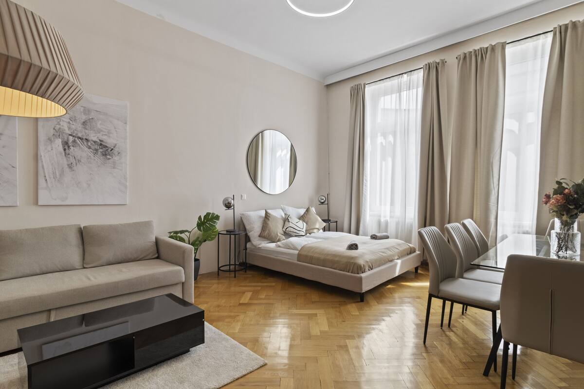 A modern living room with a sofa, a dining table with chairs, a bed, and a large window with curtains.