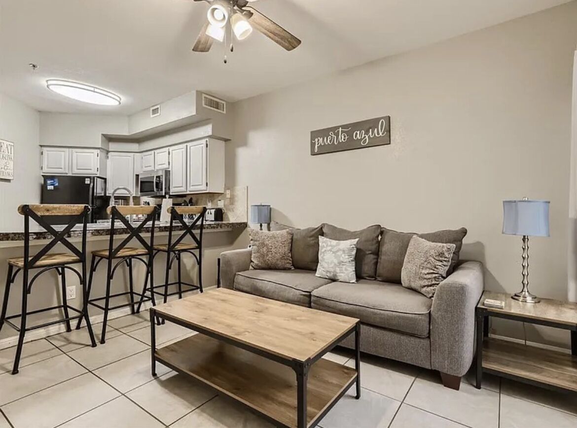 A living room with a sofa, coffee table, and bar stools. There is a kitchen area in the background with white cabinets and a microwave.