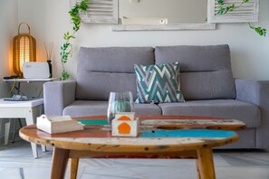 A living room with a grey sofa, a patterned throw pillow, a small table with a book and a candle, and a wall-mounted lamp.