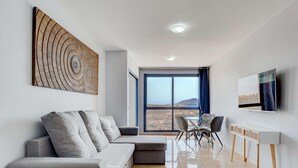 Una sala de estar moderna con un sofá modular, una mesa de comedor con sillas, un televisor montado en la pared y un amplio ventanal con vistas a un paisaje montañoso.