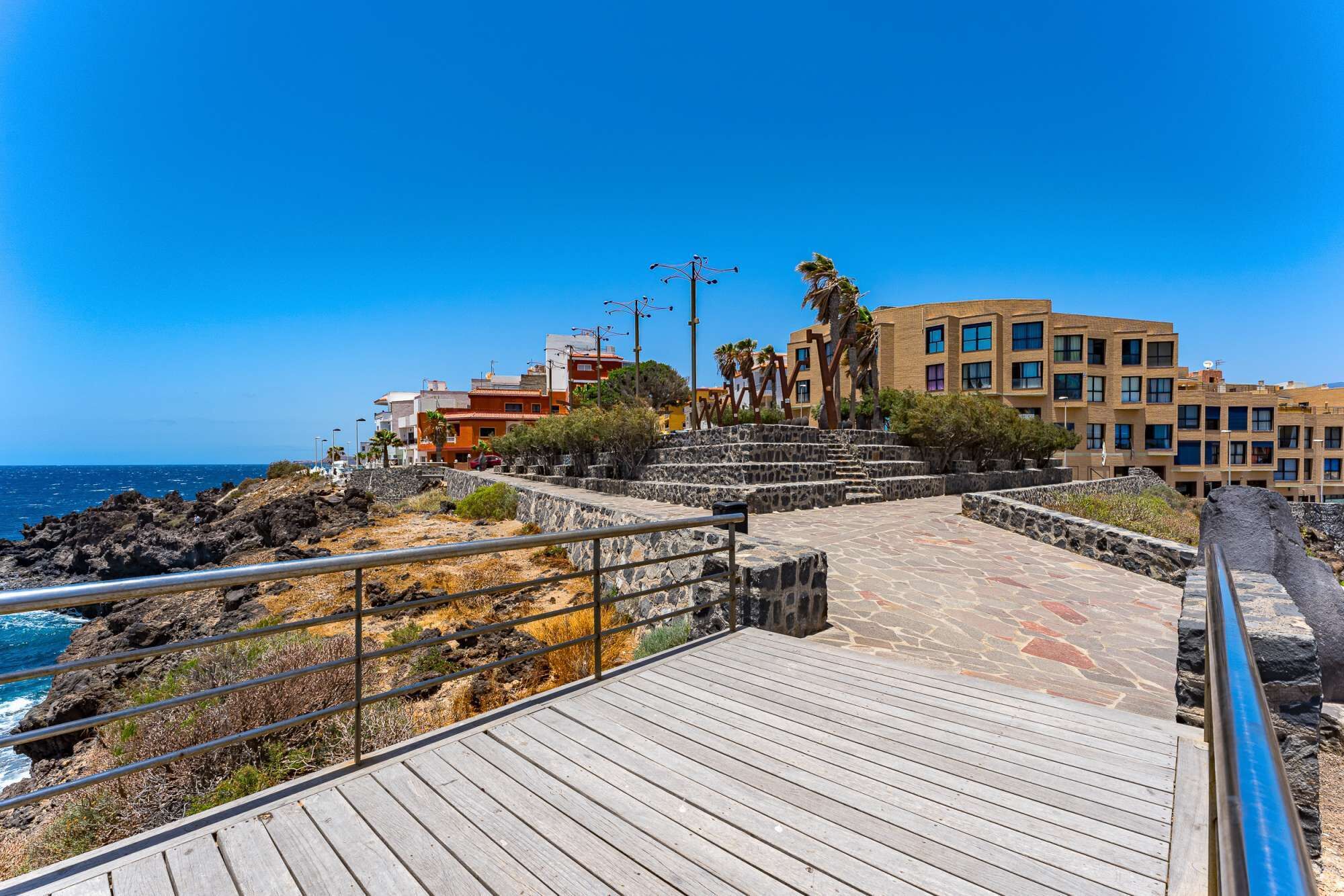 A coastal walkway with stone steps leading to a beach, surrounded by buildings and palm trees.