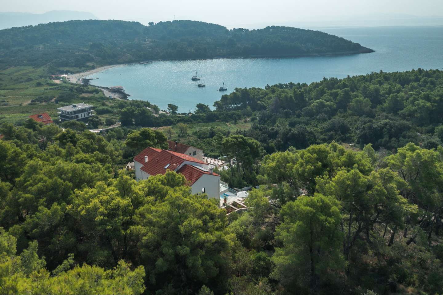 A coastal property with a large house surrounded by dense greenery, a beach, and boats in the water.