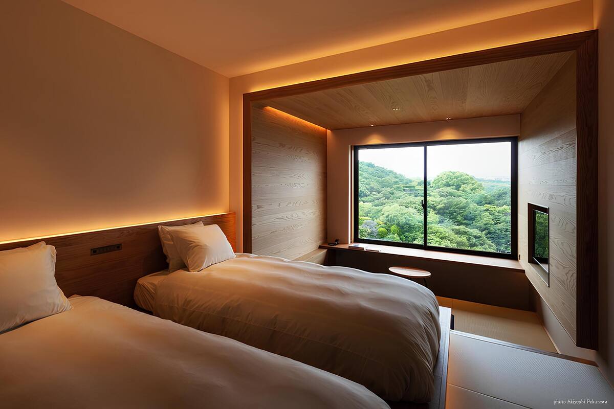 Quarto de hotel moderno com duas camas, uma janela grande com vista para uma floresta e uma mesa redonda pequena.