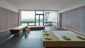 Un intérieur moderne avec un grand espace ouvert, des bancs en bois, des tatamis et une vue sur un bâtiment et la verdure environnante.
