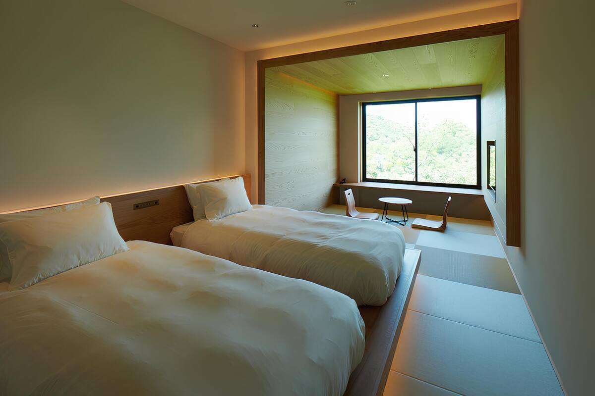 Quarto de hotel com duas camas, uma mesinha e uma janela com vista para o verde.