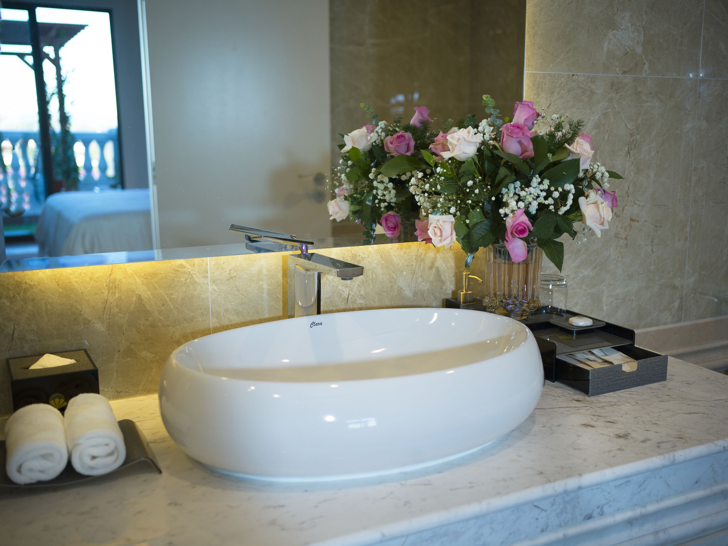 Un lavabo grande, blanco y ovalado con un grifo moderno, un espejo encima y un jarrón con flores rosas y blancas en la encimera.