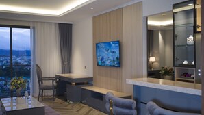 A modern hotel room with a large TV, a desk, a comfortable seating area, and a view of the city.