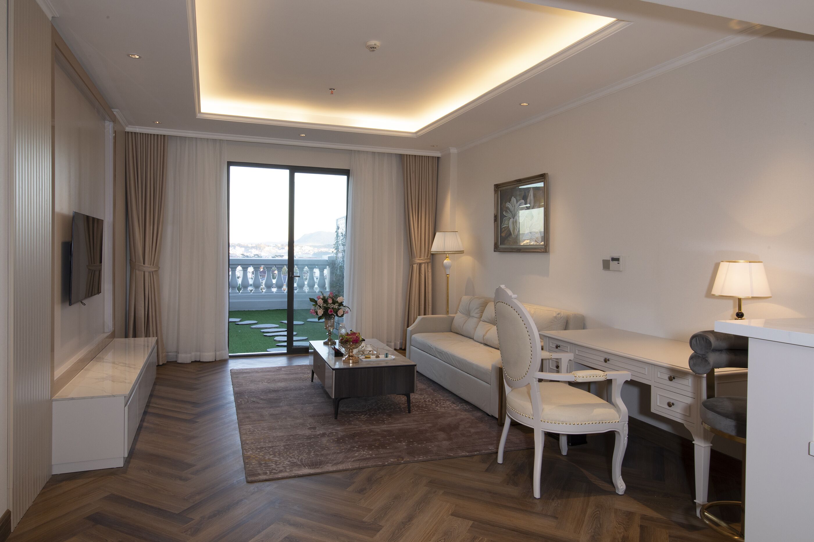 A modern hotel room with a large sliding glass door leading to a balcony, a sofa, a desk, and a chair.