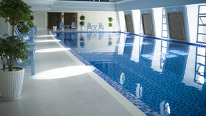 A large indoor swimming pool with clear blue water, surrounded by white walls and blue-tiled edges. There are potted plants and blue chairs along the poolside.