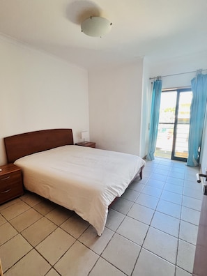 Un dormitorio con una cama grande, una mesita de noche, una lámpara y vistas a través de una ventana con cortinas.