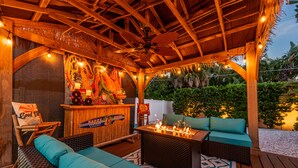 Une terrasse en bois avec un toit de chaume, des sièges d’extérieur, un bar de style tiki et des guirlandes lumineuses.