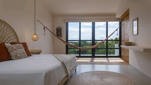 Quarto com uma cama grande, rede, tapete redondo e vista para o verde através de janelas amplas.
