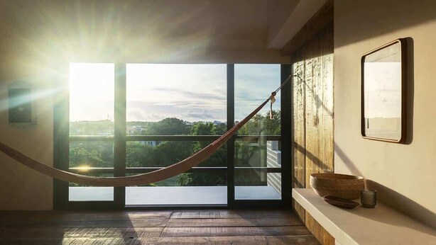 Quarto com grandes janelas, uma rede e piso de madeira.