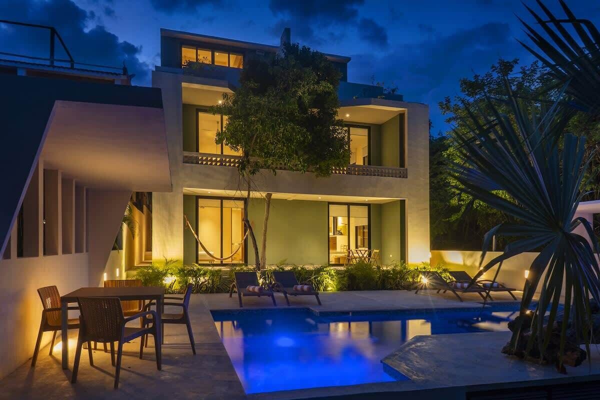 Una casa moderna de dos pisos amb piscina, mobiliari exterior i un balcó ben il·luminat.