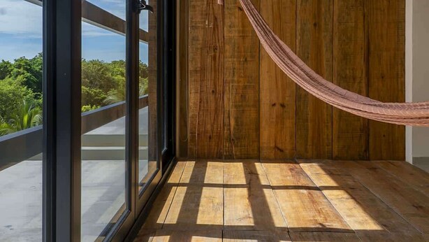 Een balkon met houten vloer, een hangmat en grote ramen met uitzicht op groen.
