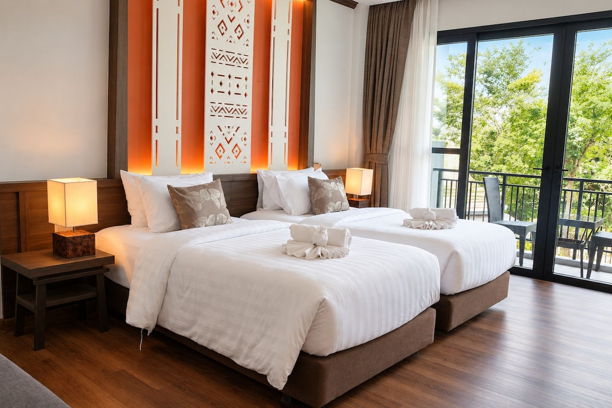 A hotel room with two beds, a nightstand with a lamp, a large window with a view of greenery, and a balcony.