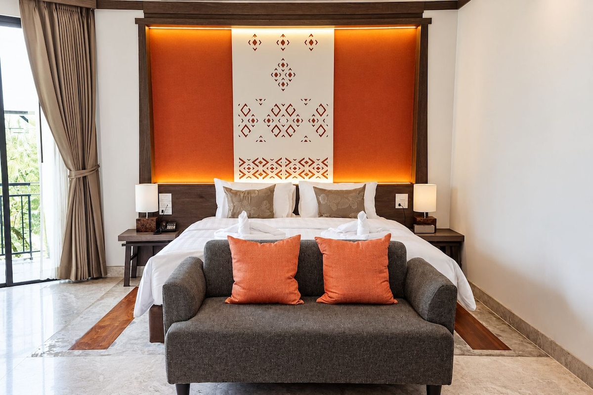 A hotel room with a large bed, two bedside tables with lamps, a grey sofa with orange cushions, and a patterned wall panel.