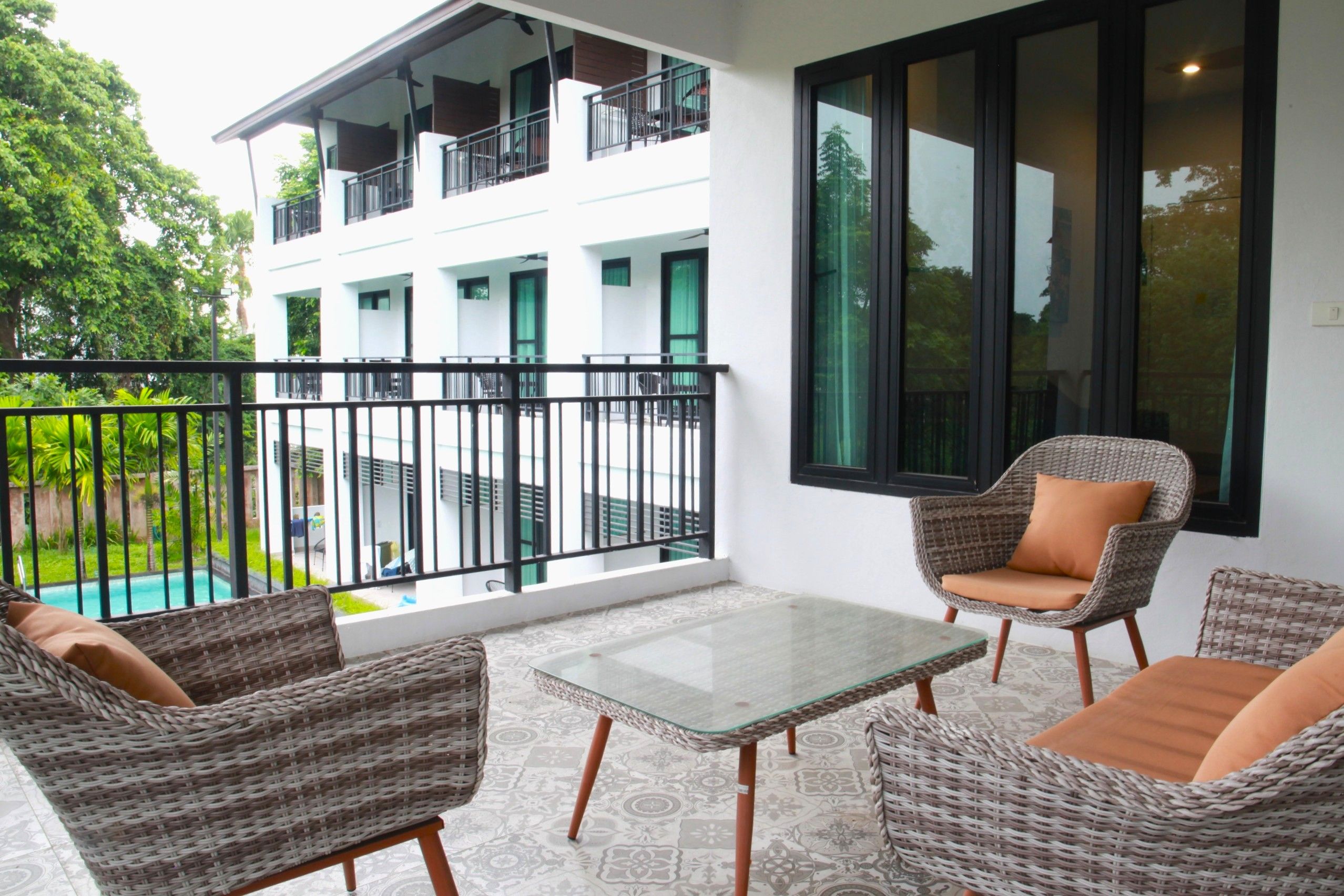 A balcony with wicker furniture, a glass table, and a view of a swimming pool and building.