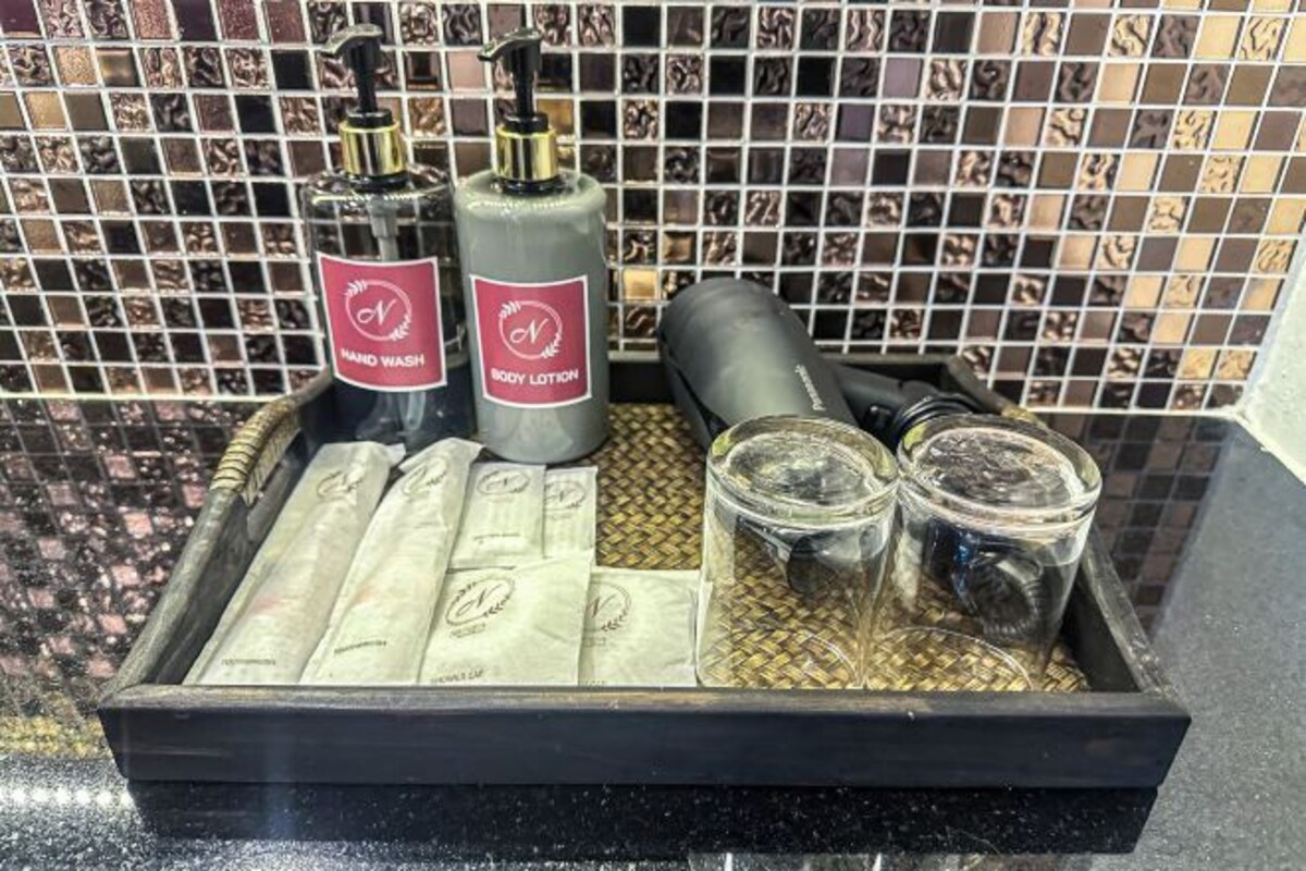 Uma bandeja com frascos de sabonete líquido e loção corporal, um objeto cilíndrico preto e dois copos de vidro com um tapete trançado por baixo.