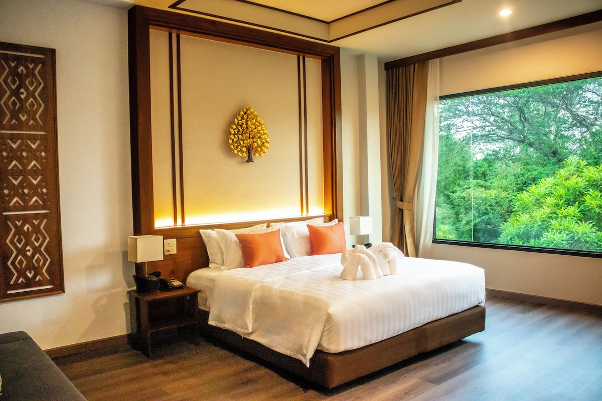 Une chambre d’hôtel avec un grand lit, une table de chevet, une lampe fixée au mur et une fenêtre donnant sur un paysage verdoyant.