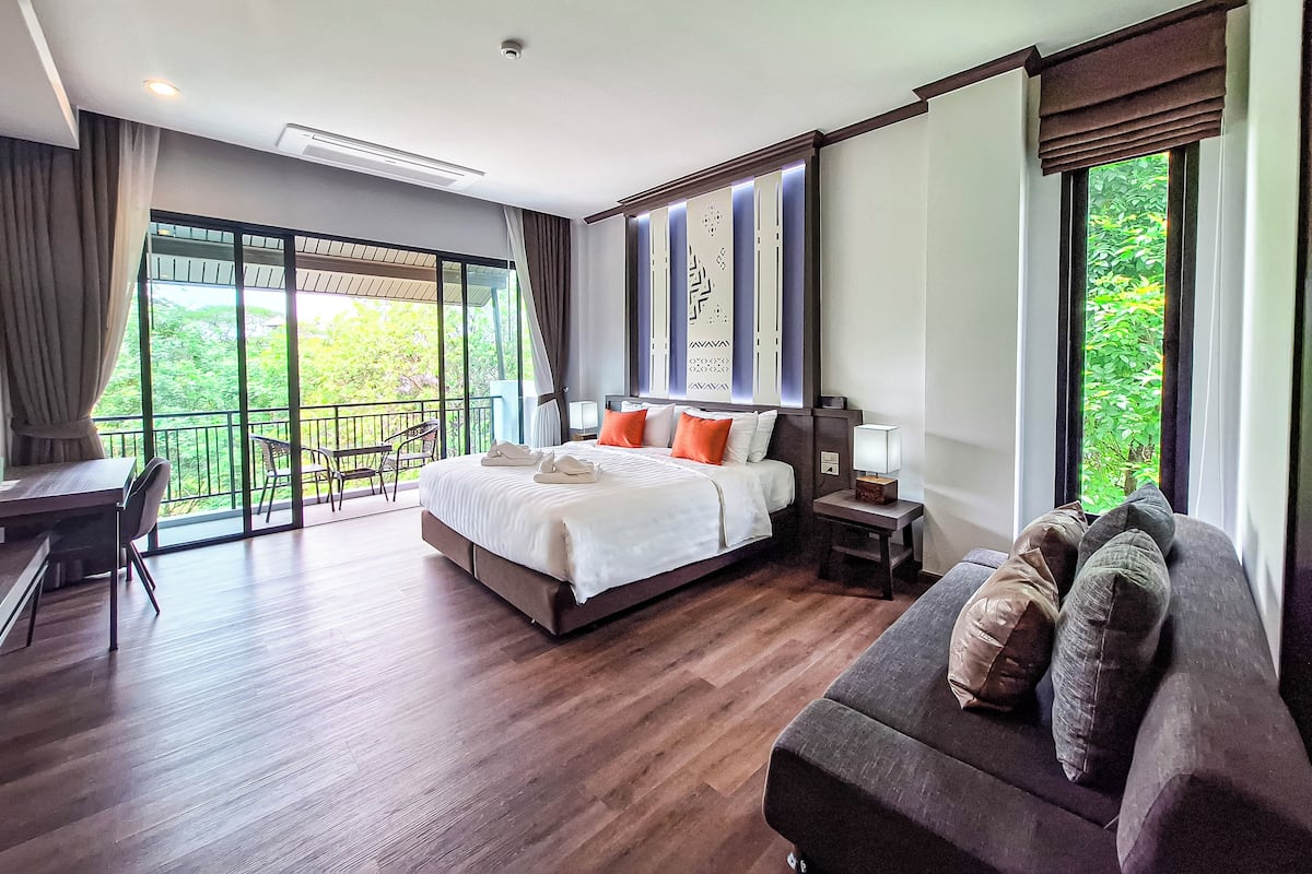 Une chambre d’hôtel moderne dotée d’un grand lit, d’un canapé, d’un bureau et d’un balcon offrant une vue sur la verdure.