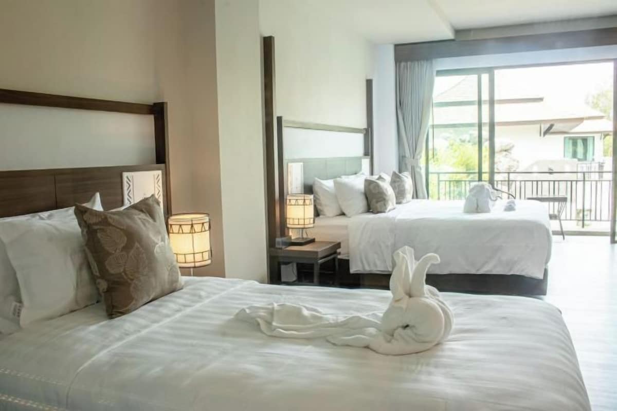 A hotel room with two beds, a nightstand with a lamp, and a view of the outside balcony.