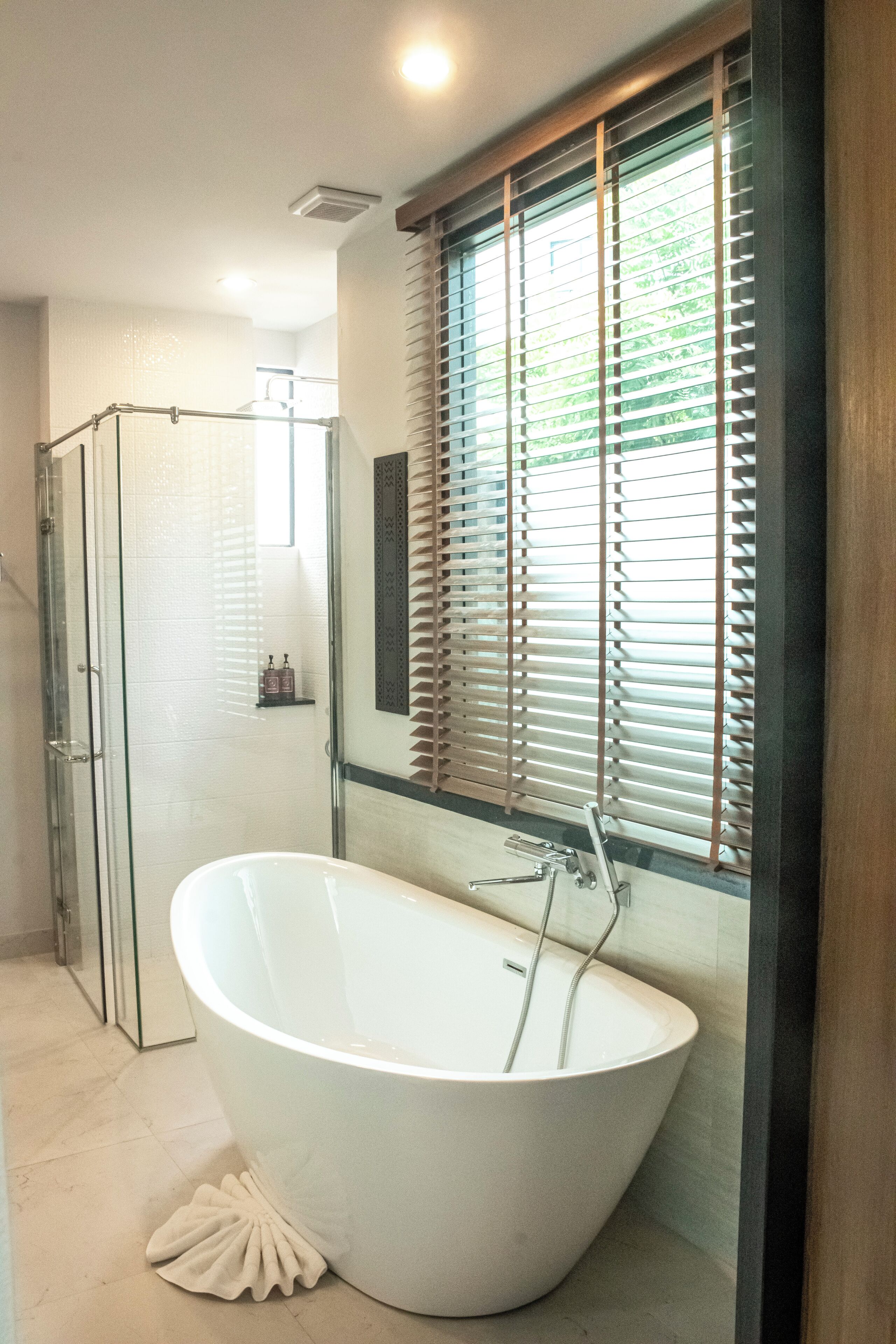 A modern bathroom with a freestanding bathtub, a glass shower enclosure, and a window with blinds.