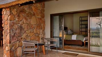 A stone wall, a glass door leading to a bedroom with a bed and a view of a tree, and two wicker chairs outside.