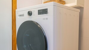 Eine weiße Waschmaschine mit Frontlader und Bedienfeld, in einem Raum mit weiß gefliesten Wänden und einer Holztür.