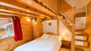 Ein Zimmer einer Holzjagdhaus mit einem Bett, einem roten Vorhang, einer an der Wand befestigten Lampe, einer Holztreppe und einem kleinen Tisch mit einer Lampe.