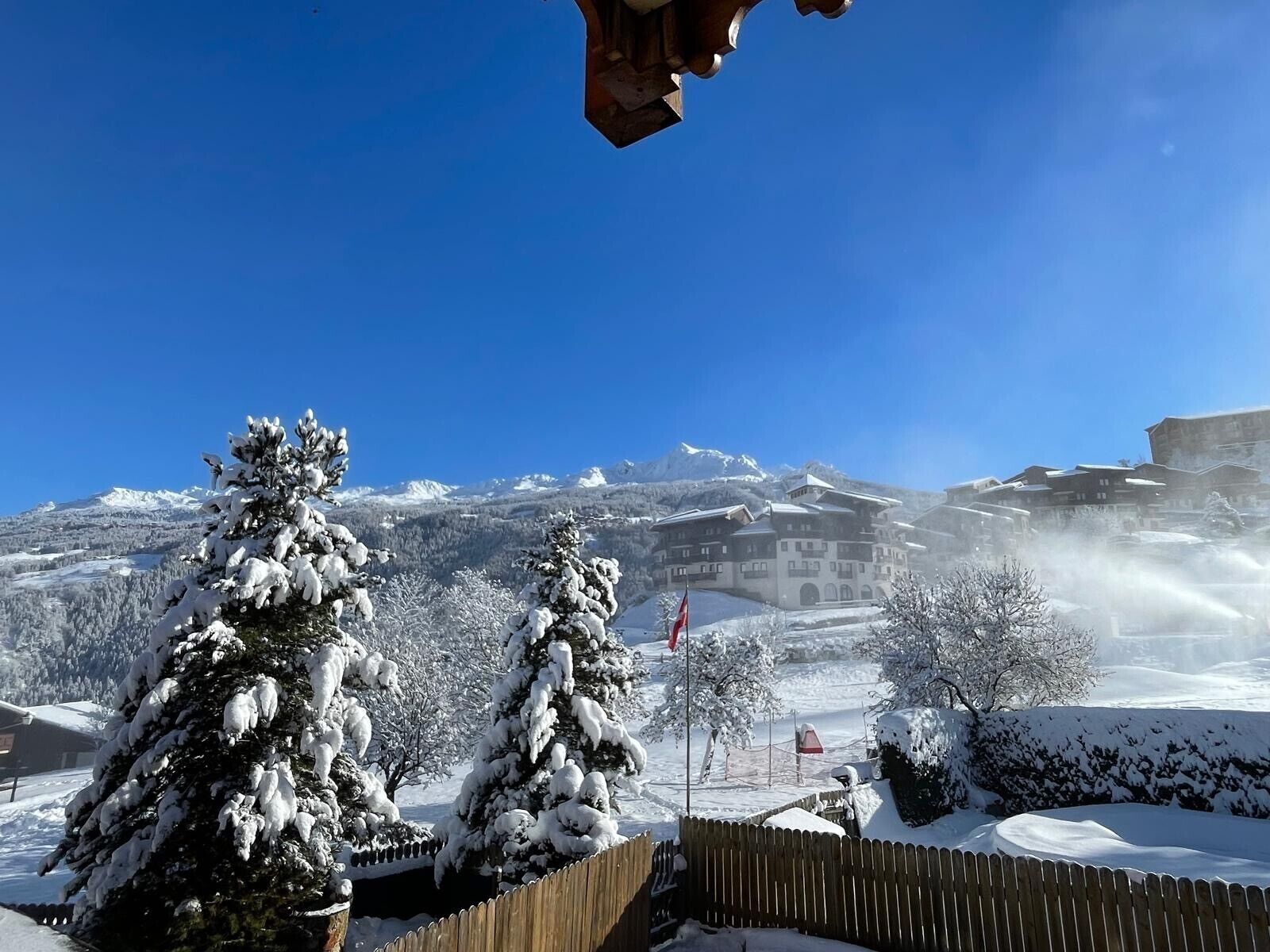 A snow-covered landscape with a building in the background, surrounded by snow-laden trees.