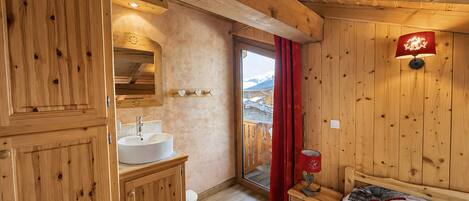 A wooden cabin interior with a wooden bed, a bedside table with a lamp, a wooden cabinet with a sink, and a window with a view of mountains.