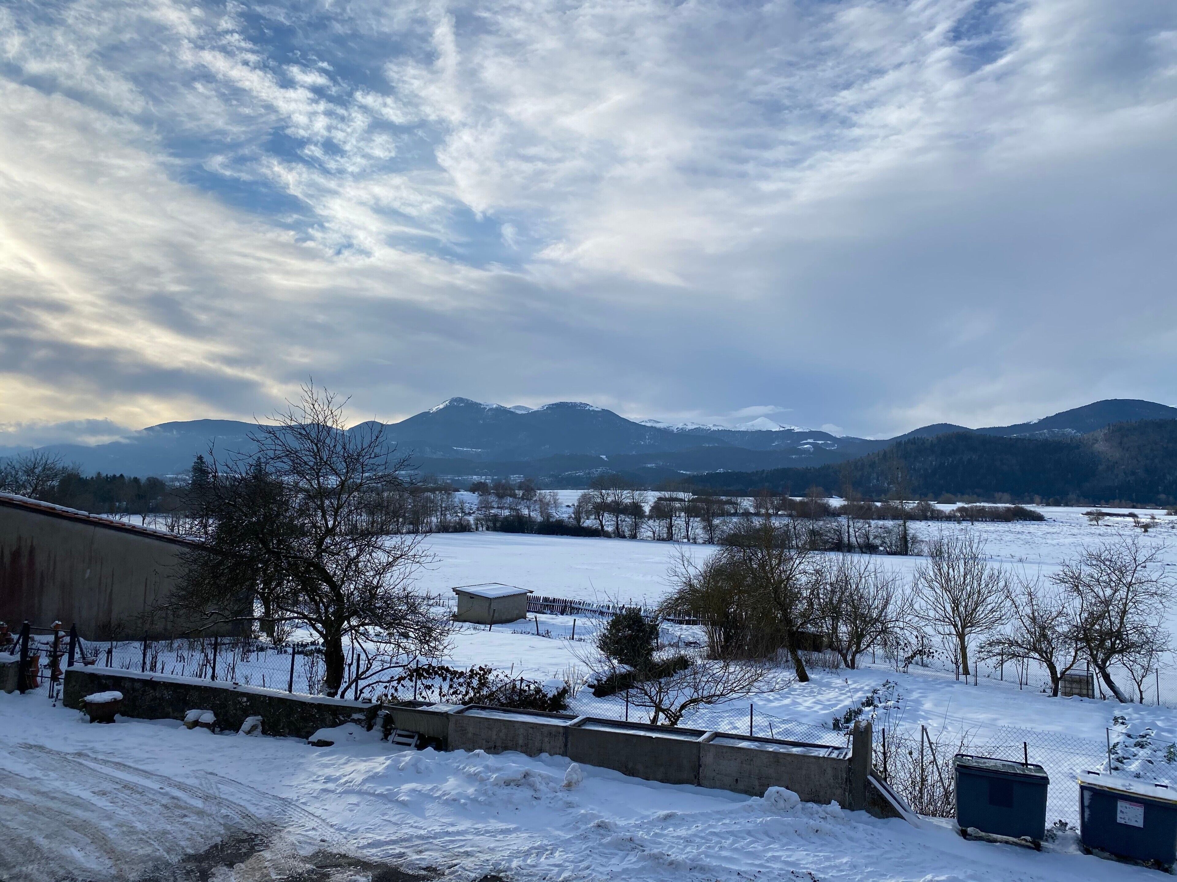 A snowy landscape with mountains in the background, a fenced area with trees, and a building in the foreground.