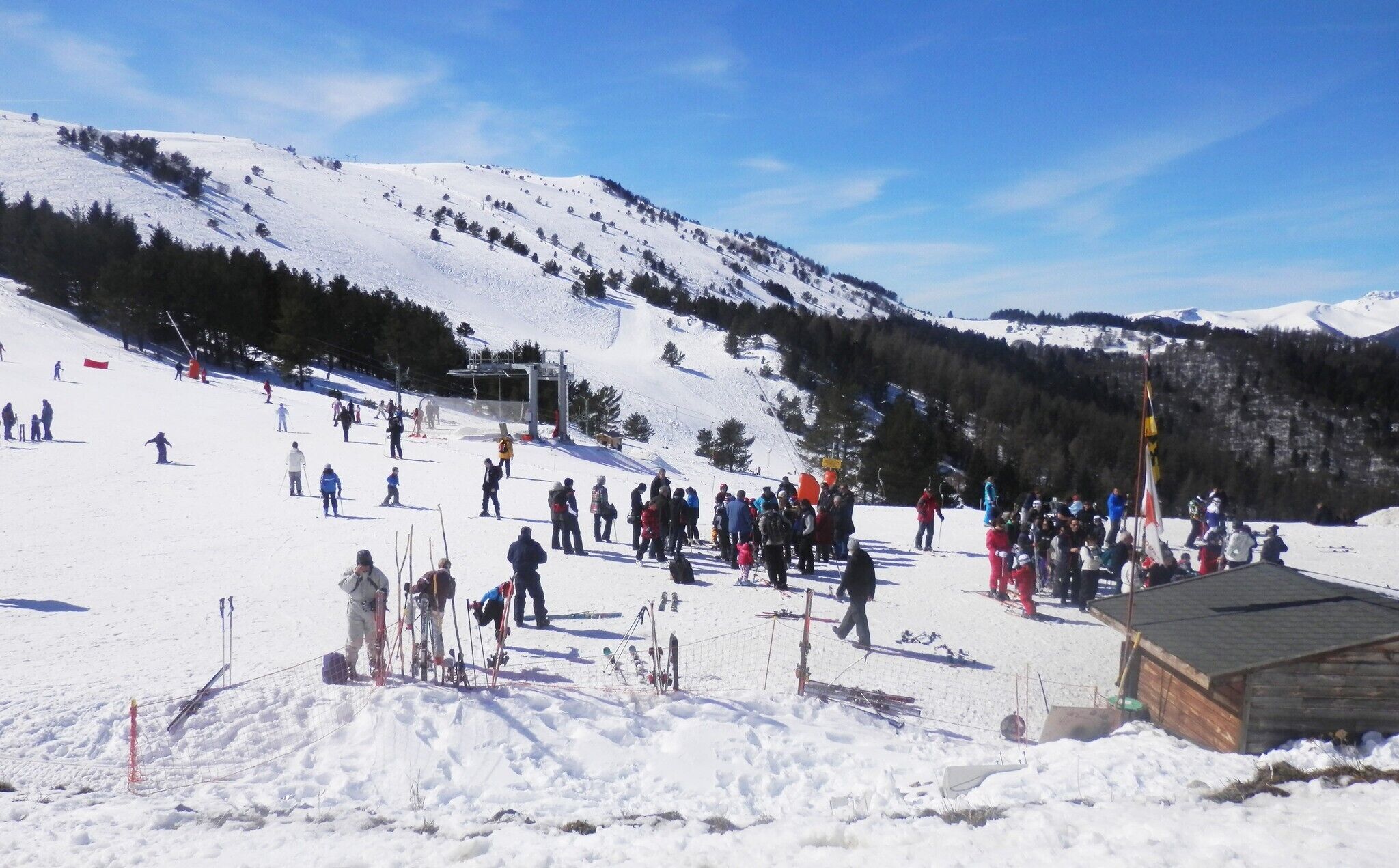 Ein verschneiter Skipisten mit Skifahrern und Snowboardern, eine Holz-Blockhütte und ein klarer Himmel.