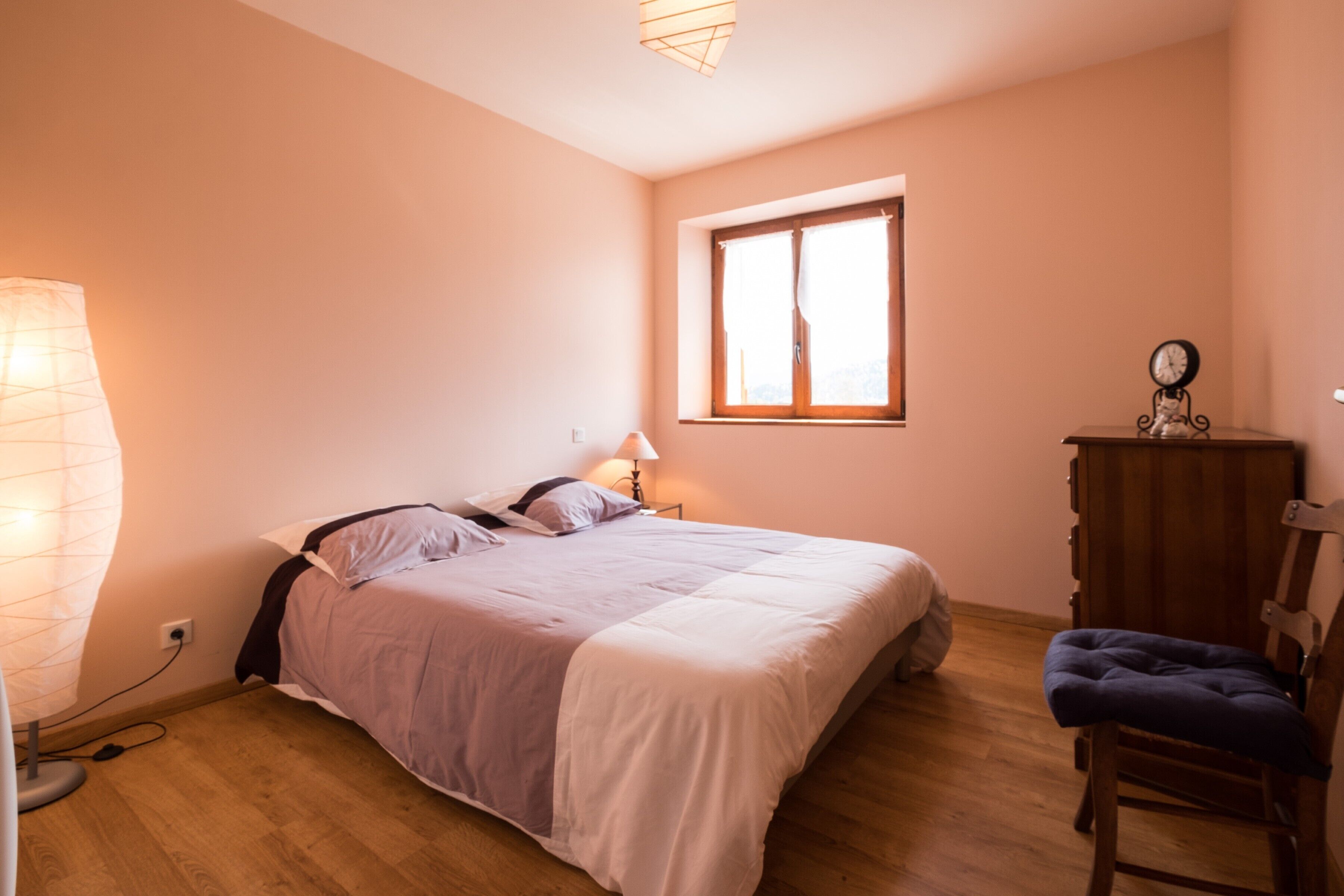 Une chambre à coucher comprenant un lit, une lampe de chevet, une commode en bois, une chaise et une fenêtre avec des rideaux.