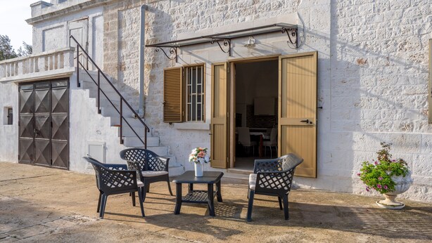 A building with a white exterior, a staircase, a door, and a small outdoor seating area with chairs and a table.